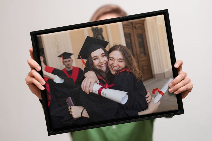 Una persona sostiene un lienzo de pared de graduación de Hofmann con dos graduados sonriendo, abrazados y sujetando sus diplomas, como inspiración para regalos de graduación personalizados e ideas de decoración mural.