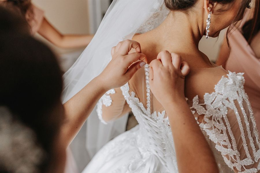 Una novia está de espaldas a la cámara mientras alguien la ayuda a abrochar su vestido de boda, capturando los últimos retoques de la checklist de fotos de la novia preparándose.