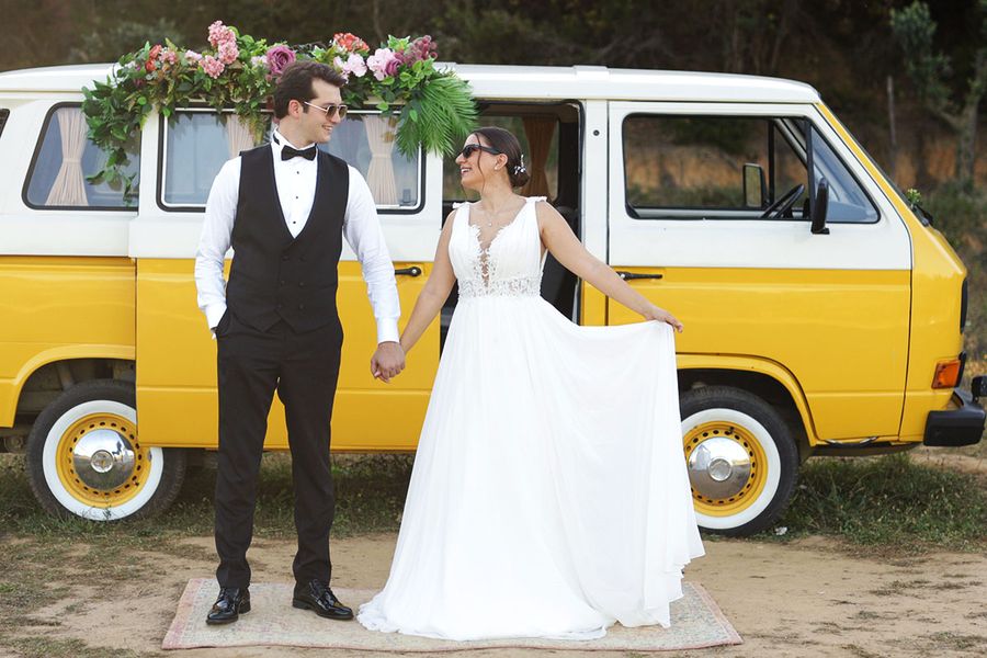 Una novia y un novio sonríen y se dan la mano delante de una furgoneta camper amarilla, capturando un momento divertido para la checklist de fotos del transporte de boda.