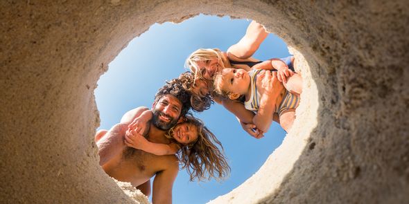 Una familia de cuatro en la playa – madre, padre, hija y bebé – mira dentro de un agujero cavado en la arena, capturando un momento divertido de verano e inspirando fotos de playa y fotografía de playa.