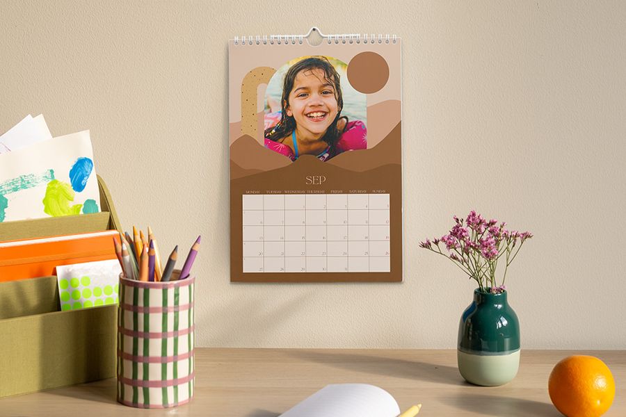 Un calendario personalizado que muestra el mes de septiembre y una foto de una niña cuelga sobre un escritorio con un portalápices, un jarrón con flores y un organizador con material de manualidades y papelería, ilustrando el consejo de Hofmann para crear un espacio de estudio enfocado para el curso escolar.