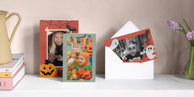 Tres tarjetas personalizadas de Halloween están colocadas una junto a otra sobre una chimenea, mostrando a una niña disfrazada de bruja, un niño disfrazado de calabaza y una niña disfrazada de fantasma rodeada de ilustraciones de fantasmas, presentando la guía de Hofmann para crear invitaciones de Halloween geniales.