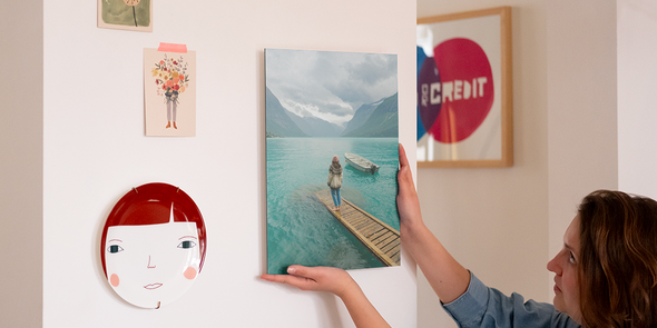 Una mujer coloca una decoración de pared personalizada en su pared, junto a un plato decorativo y algunas fotos impresas, presentando la guía paso a paso de Hofmann para colgar la decoración de pared con los kits de colgado.