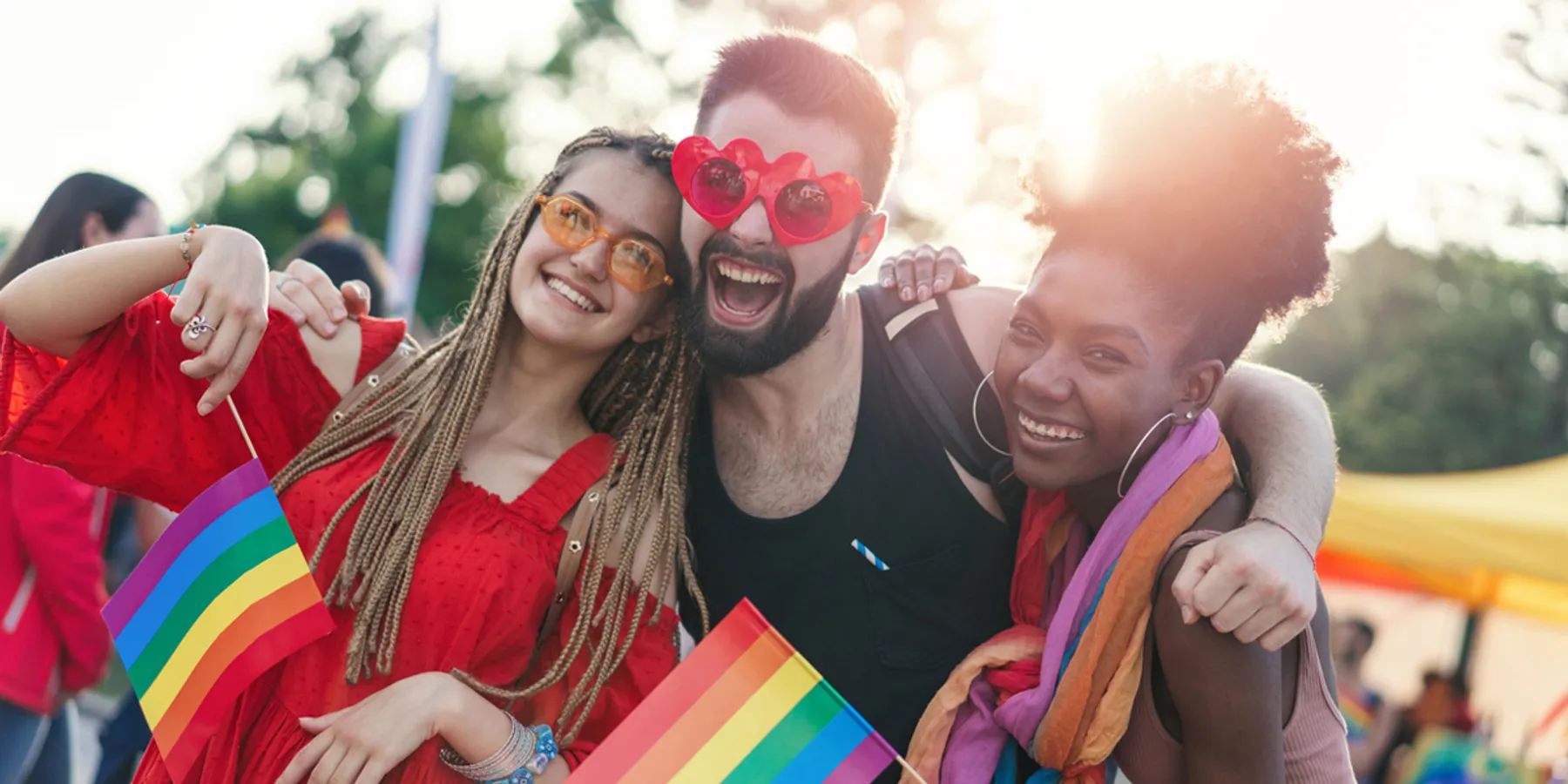 Tres amigos sonríen en un evento del Orgullo, con banderas arcoíris y gafas de sol, celebrando la alegría y la unión del Orgullo LGBTQI+.
