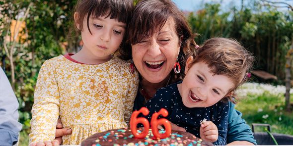 Una mujer sonríe junto a su tarta de cumpleaños con velas que muestran '66', flanqueada por una niña y un niño pequeños, presentando la guía definitiva de regalos de cumpleaños de Hofmann.