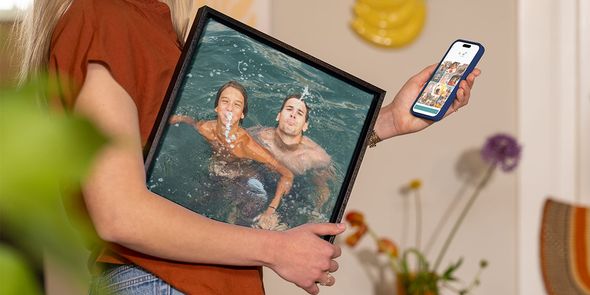 Una mujer sostiene bajo el brazo una foto de vacaciones enmarcada de un hombre y un niño pequeño jugando en el agua, mientras selecciona más fotos en su móvil, ilustrando la guía de Hofmann para convertir fotos de vacaciones en decoración de pared.