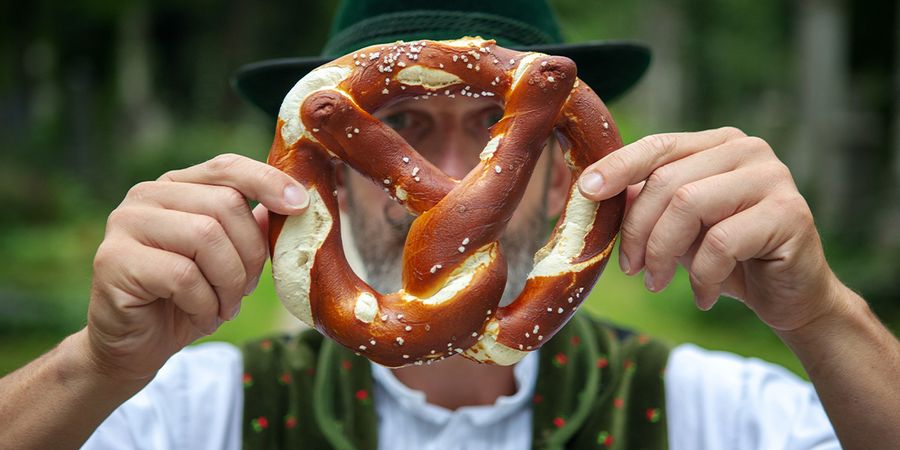 Un hombre con traje tradicional bávaro sostiene un gran pretzel frente a su cara, presentando la guía de Hofmann sobre las mejores ciudades para celebrar el Oktoberfest en Europa.