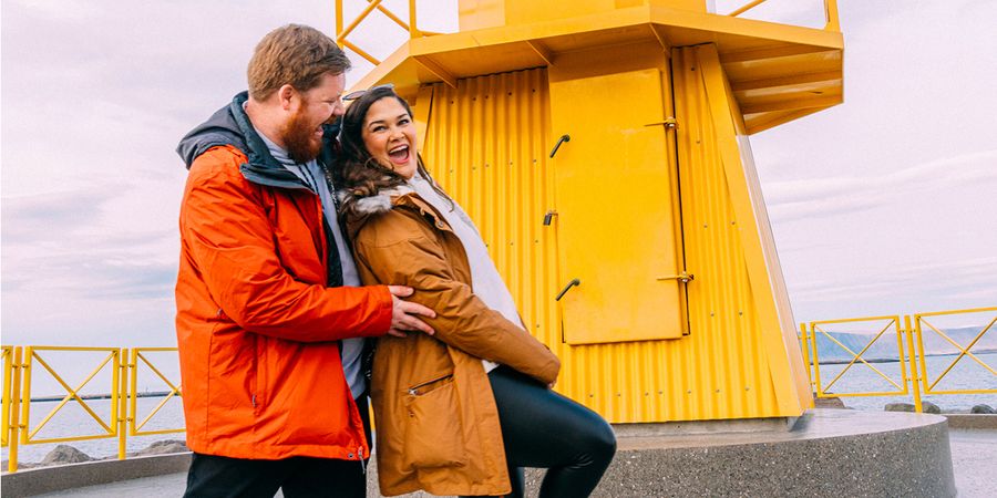 Una pareja feliz riendo frente a un faro amarillo mientras busca qué escribir en San Valentín e ideas de San Valentín para sus fotos y regalos de Hofmann.