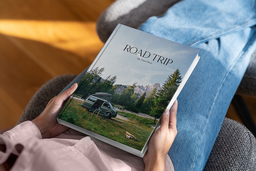 Un coffee table book de Hofmann titulado Road Trip reposa sobre el regazo de una persona, con un paisaje de bosque y una autocaravana en la portada, como inspiración para un álbum decorativo de viajes.