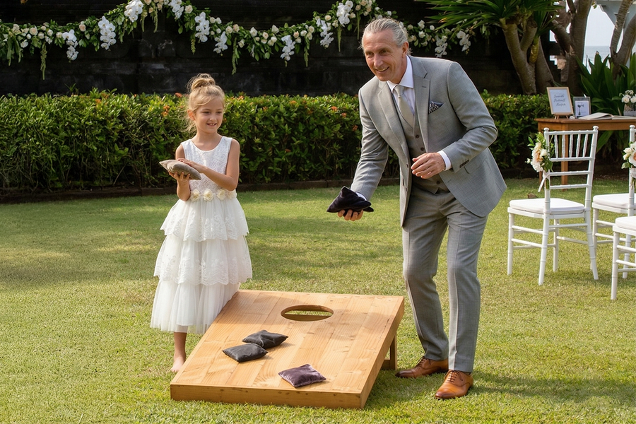 Una niña vestida de gala y un hombre mayor con traje juegan al cornhole en una boda, lanzando saquitos al tablero, como inspiración para Hofmann y para juegos de boda al aire libre y actividades para invitados.
