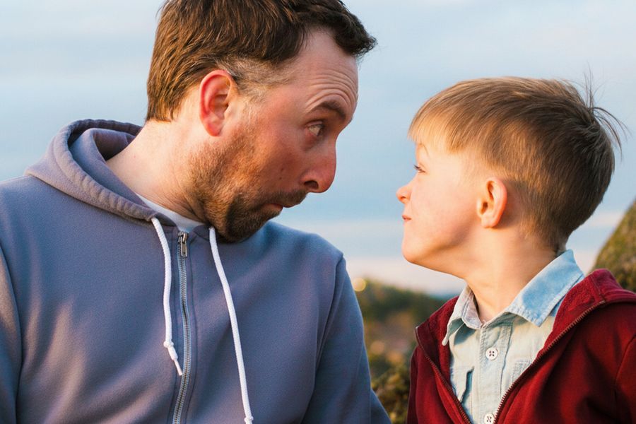 Un padre y su hijo ponen caras divertidas mientras se miran, como inspiración para una tarjeta Día del Padre divertida y frases Día del Padre con humor.