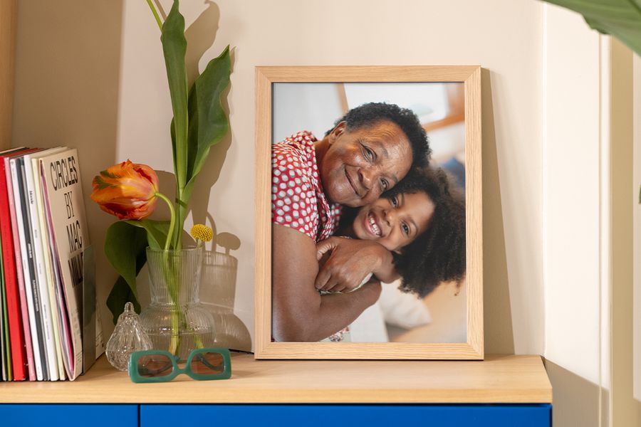 Una foto enmarcada de una abuela y su nieta abrazándose está sobre una mesa junto a libros, una revista y un jarrón original, como inspiración para tendencias en marcos y formas asequibles de realzar la decoración de pared.