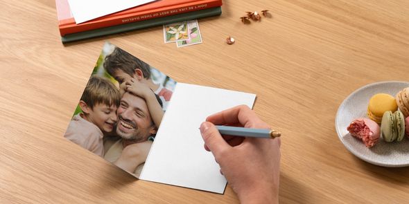 Una tarjeta de fotos personalizada de Hofmann para el Día del Padre abierta sobre una mesa muestra a un padre con sus dos hijos, con un bolígrafo listo para escribir un mensaje junto a unos libros y un plato de macarons, como inspiración para qué escribir en una tarjeta del Día del Padre.