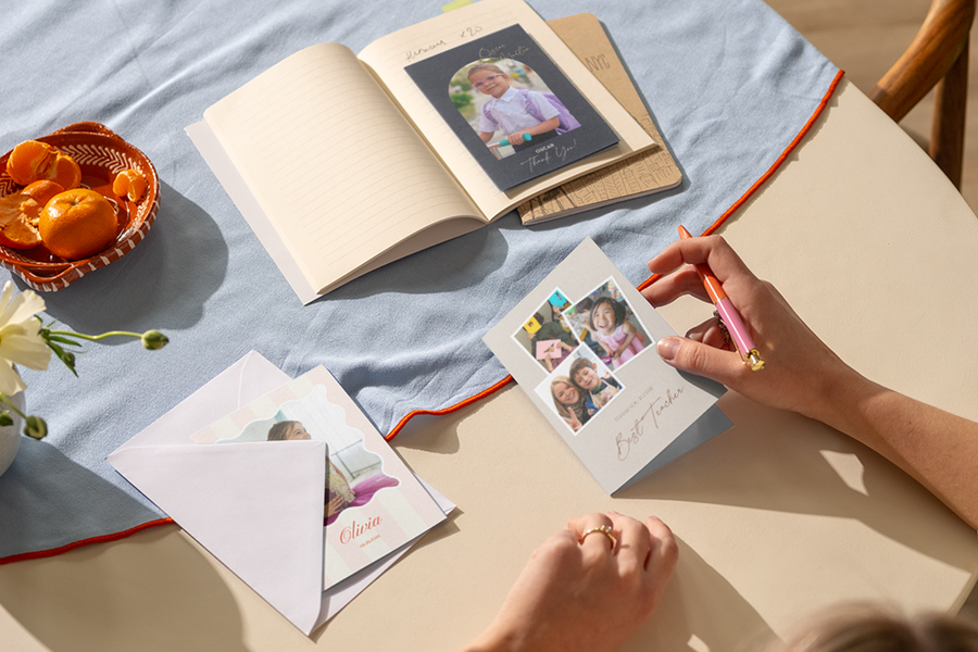 La mano de una mujer descansa sobre una mesa, lista para escribir en una tarjeta de agradecimiento para profesor personalizada de Hofmann con varias fotos de niños felices.
