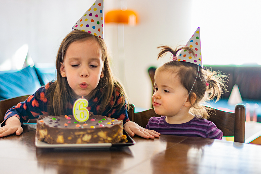Una niña con gorro de fiesta soplando la vela de su tarta de 6º cumpleaños junto a una amiga, una idea perfecta de regalos de cumpleaños para niños de Hofmann.
