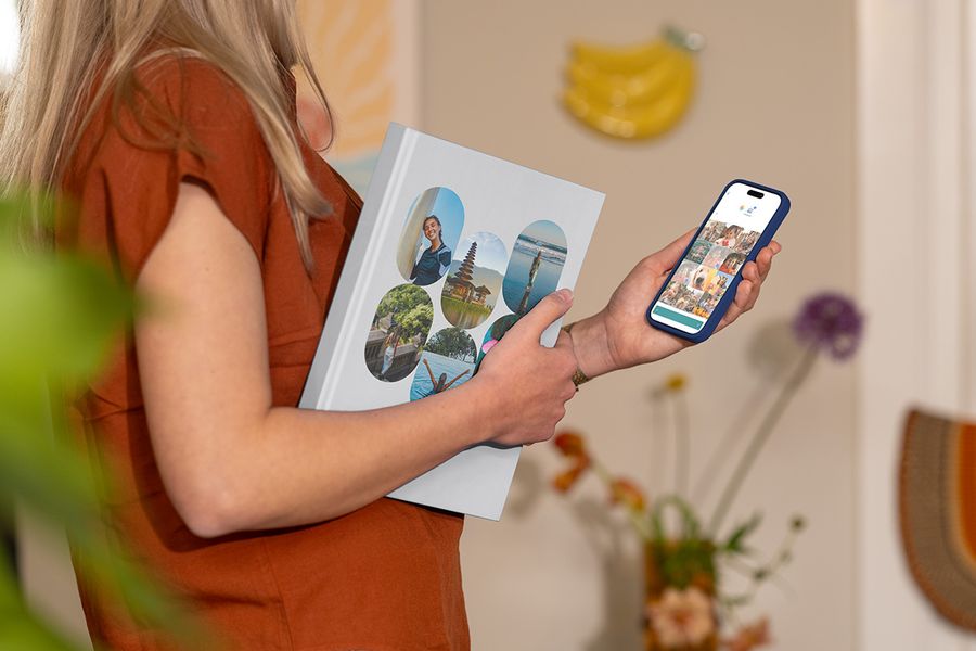 Una mujer sostiene un álbum de vacaciones de Hofmann cerrado con fotos familiares de viaje en la portada mientras mira imágenes de viaje en su móvil, ilustrando cómo elegir las mejores fotos para un álbum de fotos de viaje personalizado.