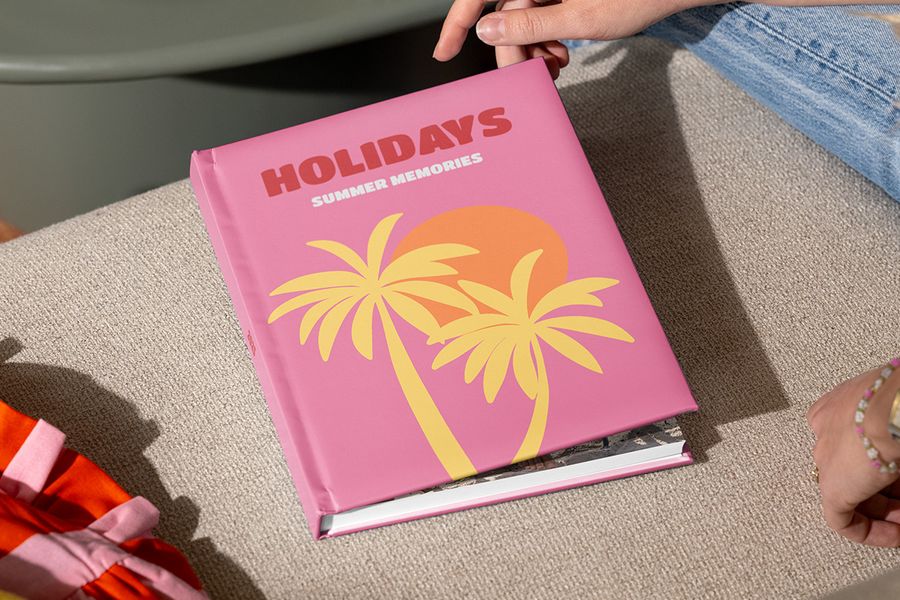 Un álbum de viajes de Hofmann cerrado, titulado “Holidays, summer memories” y con palmeras ilustradas sobre una portada rosa, es sostenido por unas manos listas para abrirlo, ilustrando cómo estructurar un álbum de fotos de viaje y contar la historia del viaje.