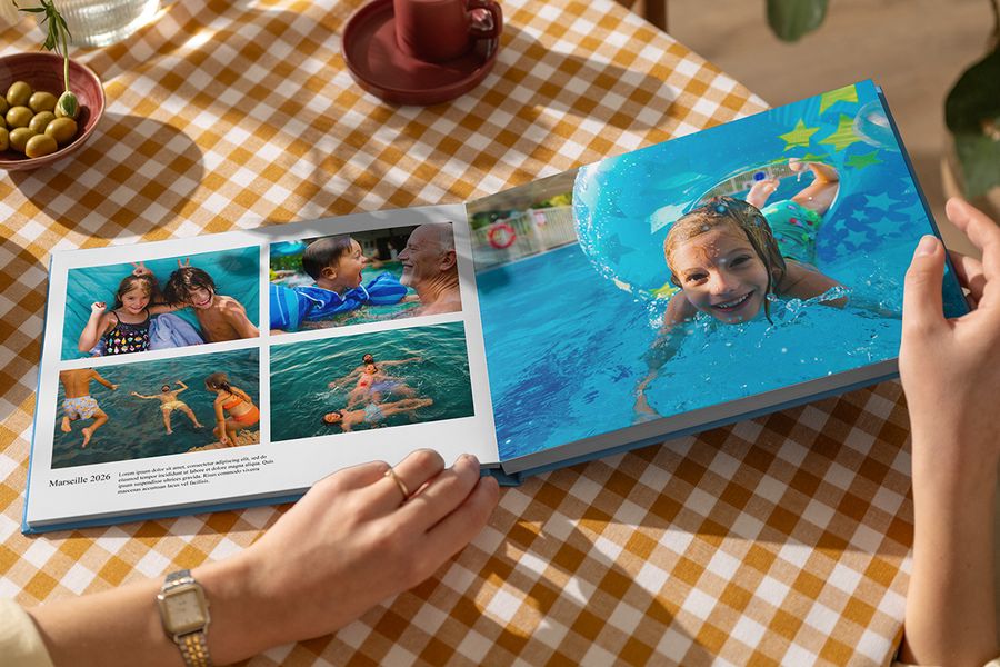 Un álbum de vacaciones de Hofmann abierto muestra fotos familiares de verano jugando en el agua, con una página completa y otra titulada Marseille 2026 con texto y descripción, ilustrando cómo los pies de foto dan vida a un álbum de fotos de viaje personalizado.