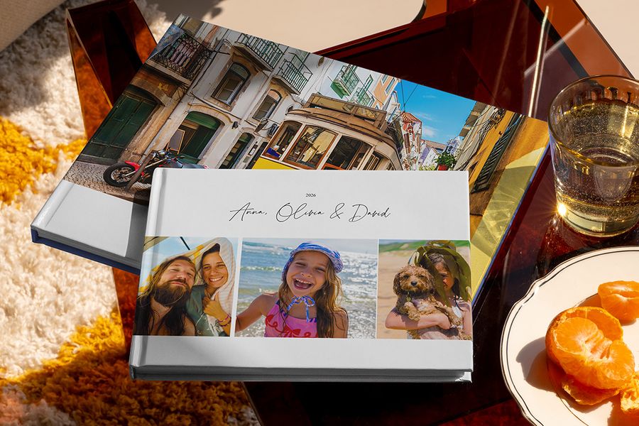Se muestran juntos dos fotolibros de viaje de Hofmann, uno en formato horizontal con fotos familiares de viaje en la portada y otro con una imagen de un tranvía de vacaciones, como inspiración para distintos tipos de álbum de fotos de viaje personalizado.
