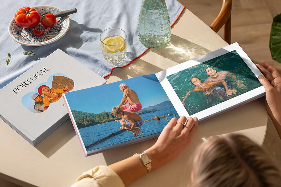 Se muestran dos álbumes de viaje de Hofmann, mientras una mujer hojea un álbum de fotos de viaje abierto con fotos familiares junto al mar, al lado de un álbum cerrado de Portugal, ilustrando un álbum de vacaciones personalizado lleno de recuerdos de viaje.