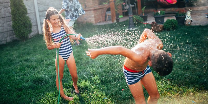 Una niña y un niño juegan en el jardín con una manguera, un momento divertido de vacaciones en casa de la guía de Hofmann para planificar las mejores vacaciones en casa.