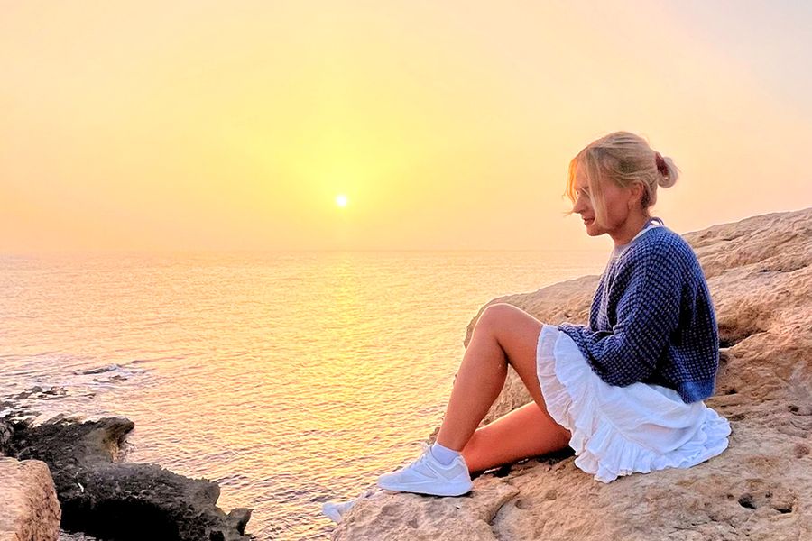 Una mujer está sentada sobre unas rocas mirando el mar y la puesta de sol, reflejando el espíritu de las ideas de vacaciones en casa de aventura de Hofmann.