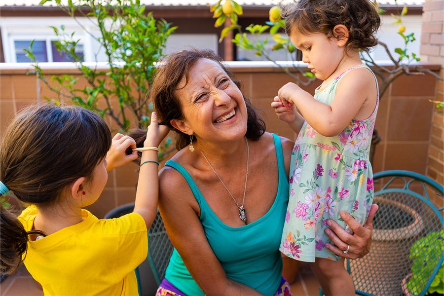 Una mujer está sentada en un balcón mientras dos niñas juegan con su pelo y la peinan, un momento divertido de las ideas de vacaciones en casa en familia de Hofmann.