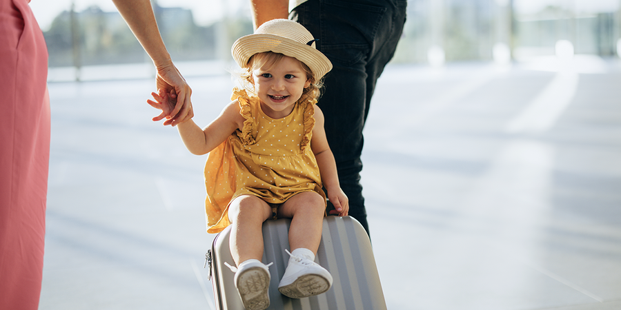 Una niña pequeña va sentada sobre una maleta mientras la arrastran, capturando un momento divertido de viajes en familia y de viajar con niños con Hofmann.