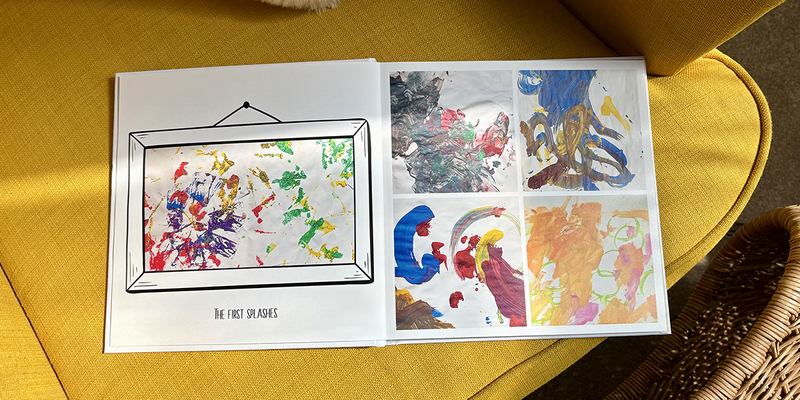 Un álbum de fotos infantil abierto descansa sobre un sillón amarillo, con las primeras pinceladas de pintura de un niño pequeño en la página izquierda y cuatro pinturas diferentes en la derecha, ilustrando las ideas de Hofmann para un álbum de fotos infantil.