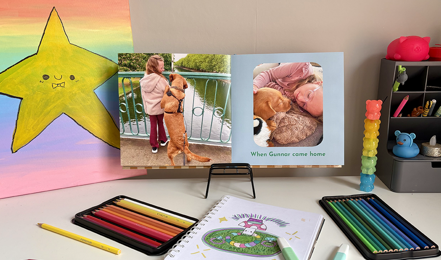 Un álbum de fotos infantil abierto descansa sobre el escritorio de un niño, rodeado de lápices de colores y dibujos, con una foto de una niña y su perro en la página izquierda y otra imagen con pie de foto en la derecha, ilustrando los consejos de Hofmann para combinar fotos y obras de arte en un álbum de fotos familiar.