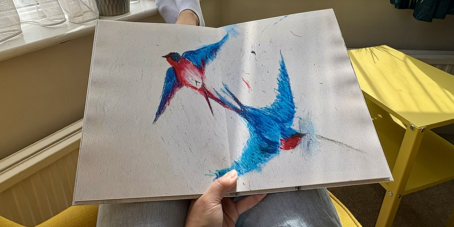 Un álbum de fotos infantil abierto con una doble página de un dibujo de pájaros descansa sobre el regazo de una mujer sentada en un sillón, ilustrando los pasos de Hofmann para crear un álbum de fotos familiar a partir de los dibujos de su hijo.