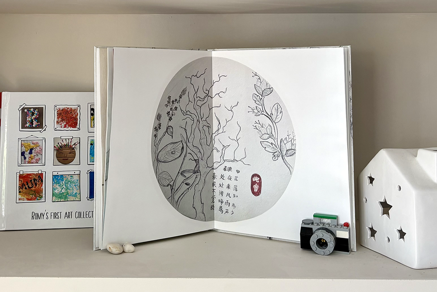 Un álbum de fotos infantil abierto muestra una doble página de bocetos de árboles con texto, junto a un álbum cerrado con una portada estilo scrapbook que reúne varios ejemplos de obras de arte infantiles, ilustrando el recuerdo significativo que crea un álbum de fotos infantil de Hofmann.