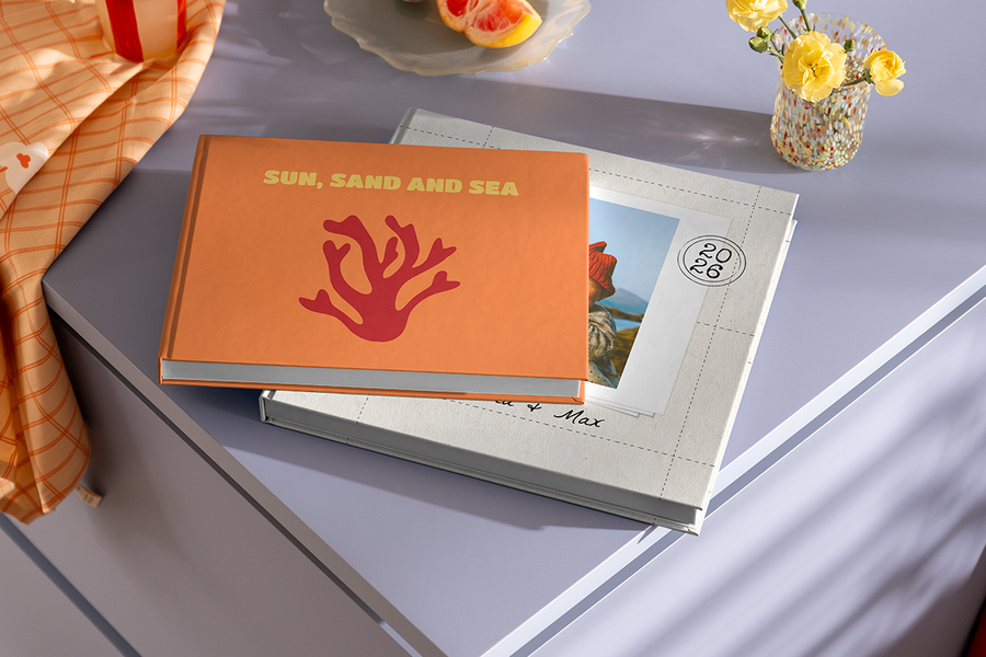Dos álbumes de fotos están apilados uno sobre otro, con el álbum de arriba luciendo un diseño de coral y el título 'Sun, Sand and Sea', ilustrando las ideas de título para álbumes de fotos de viajes a la playa de Hofmann.