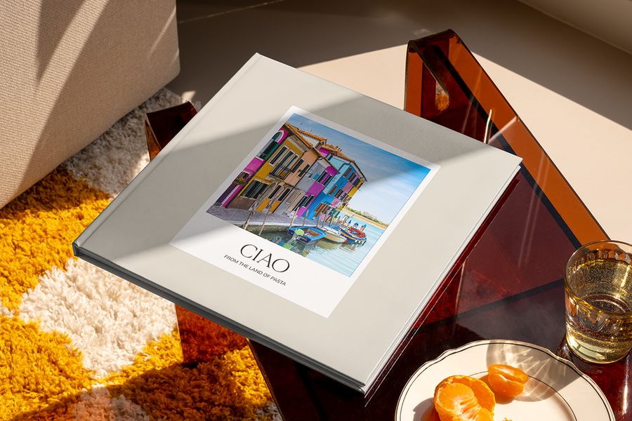 Un álbum de fotos con un canal y casas de colores en Italia en la portada se titula 'Ciao from the Land of Pasta', ilustrando las ideas de título para álbumes de fotos de viajes a ciudades de Hofmann.