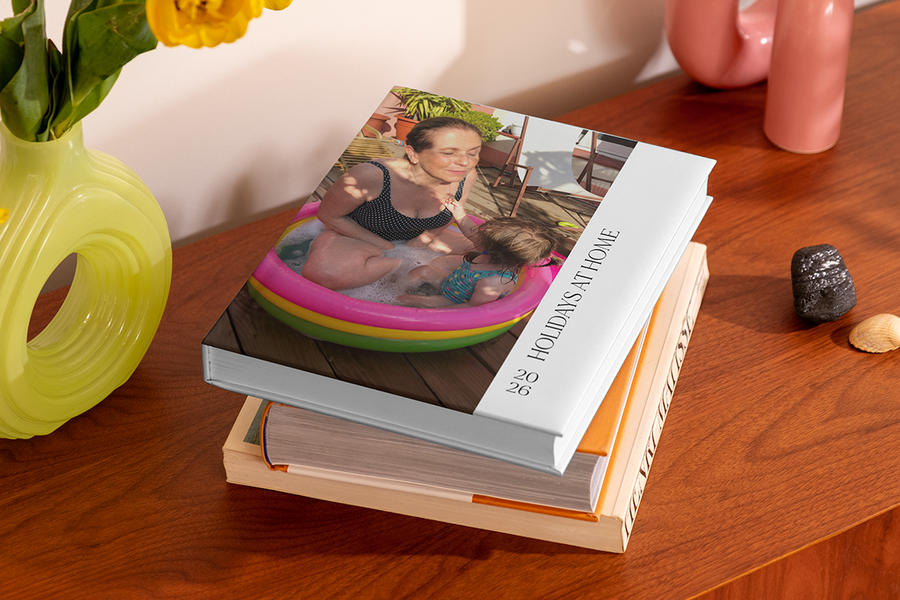 Una pila de álbumes de fotos tiene arriba un álbum que muestra a una mujer y un niño pequeño en una piscina hinchable, titulado 'Holidays at Home', ilustrando las ideas de título para álbumes de fotos de vacaciones en casa de Hofmann.