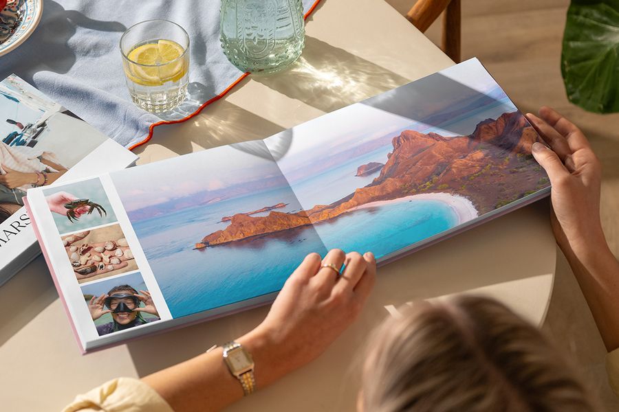 Un álbum de fotos de viaje abierto sobre una mesa muestra una doble página con un precioso paisaje de playa junto a tres fotos de verano, ilustrando el diseño Travel Chapter de la nueva Summer Travel Series de Hofmann.
