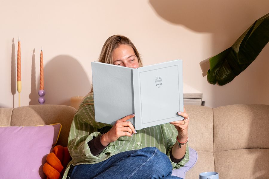 Una mujer está sentada en el sofá junto a un cojín de colores, mirando un álbum de fotos de viaje personalizado titulado 'Oslo', que muestra los nuevos diseños de la Summer Travel Series de Hofmann.