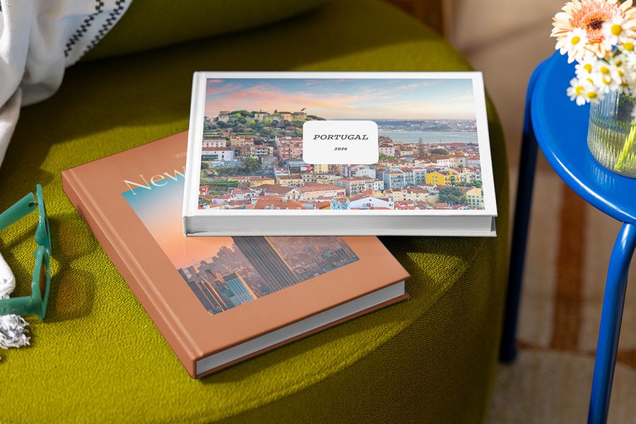 Dos álbumes de fotos personalizados están apilados sobre un taburete, el de arriba titulado 'Portugal' con una imagen de Portugal, y el de abajo titulado 'New York' con una imagen de la ciudad, mostrando los diseños personalizables de la Summer Travel Series de Hofmann.