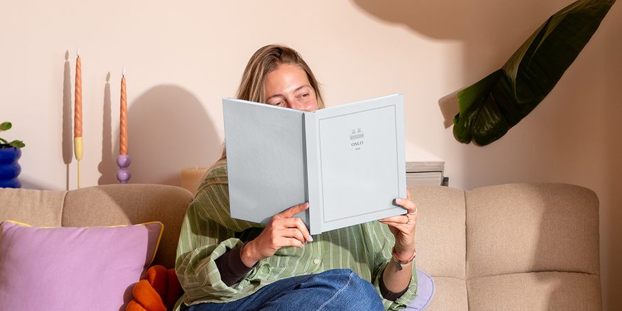Una mujer está sentada en el sofá junto a un cojín de colores, mirando un álbum de fotos de viaje personalizado titulado 'Oslo', que muestra los nuevos diseños de la Summer Travel Series de Hofmann.