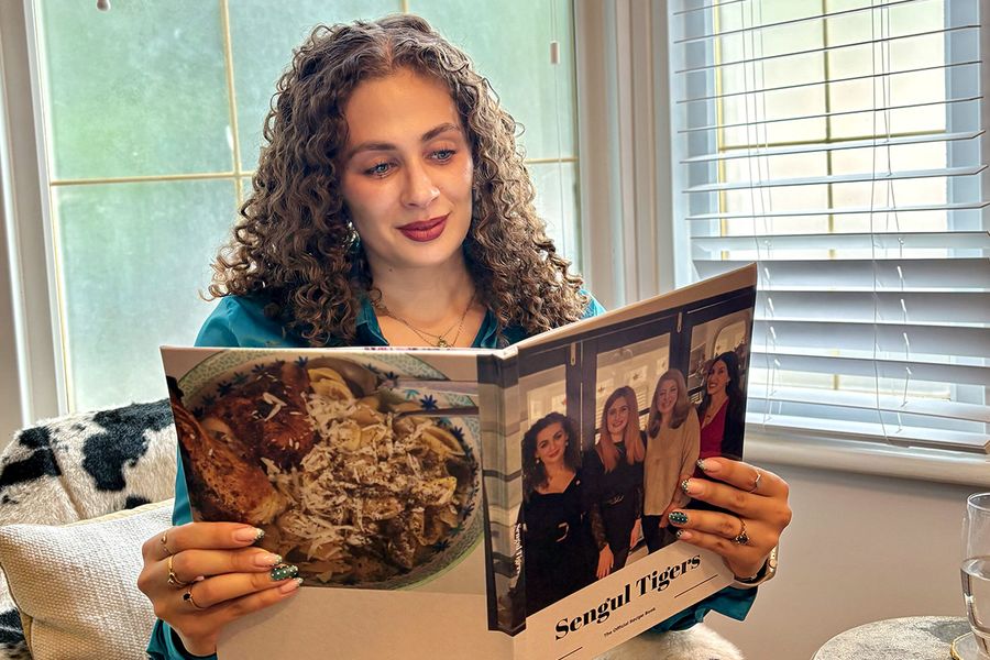 Sarah, que trabaja en Hofmann, está sentada en una silla con estampado de vaca mirando su libro de recetas familiar personalizado, cuya portada muestra a cuatro mujeres, entre ellas Sarah, ilustrando cómo un libro de recetas personalizado reúne recetas y recuerdos familiares.