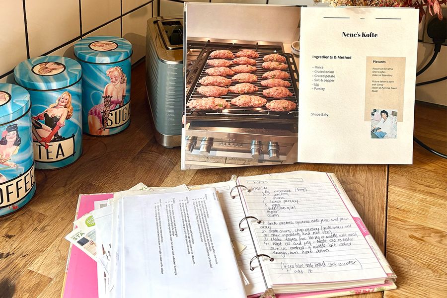 El libro de recetas personalizado abierto de Sarah muestra una receta de kofte de su Nene, colocado frente a una vieja carpeta abierta con recetas familiares manuscritas, ambos sobre una encimera de cocina, ilustrando cómo reunió recetas de varias generaciones en un solo libro.