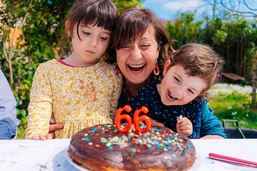 Una mujer sonríe junto a su tarta de cumpleaños con velas que muestran '66', flanqueada por dos niños sonrientes, ilustrando los consejos de Hofmann para captar momentos de cumpleaños multigeneracionales.