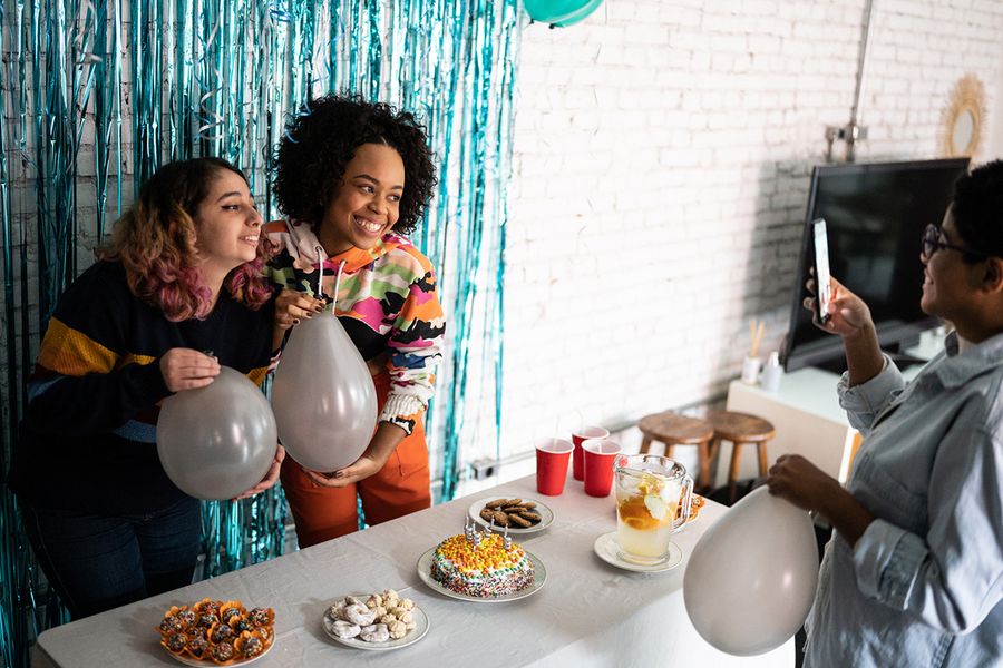 Dos niñas posan frente a un fondo de flecos brillantes, sosteniendo globos junto a una mesa de aperitivos llena de comida de fiesta y una tarta de cumpleaños, ilustrando los consejos de Hofmann para fotografiar la decoración y las mesas de comida de la fiesta.