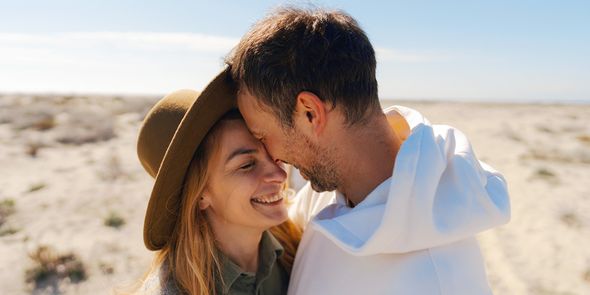 Una pareja en la treintena sonríe y se abraza en un entorno de playa desértica, presentando la guía de Hofmann con 60 ideas de mensajes para una tarjeta de aniversario de bodas.