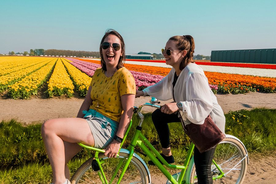 Dos mujeres montan en bicicleta juntas, sonriendo y riendo en un campo lleno de flores, ilustrando las ideas de Hofmann sobre qué escribir en una tarjeta de aniversario de bodas para amigos.