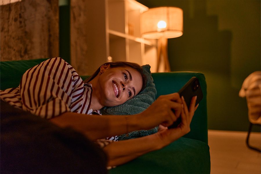 Una mujer está tumbada en un sofá verde oscuro mirando su móvil en un salón, iluminada por una lámpara que crea una luz acogedora, ilustrando los consejos de Hofmann para una iluminación hygge en casa.
