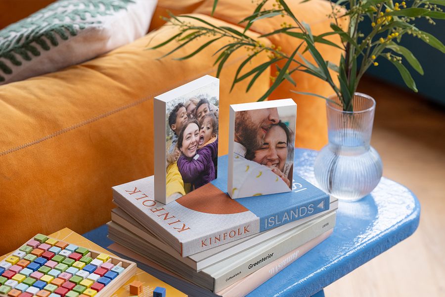 Dos bloques con foto personalizados descansan sobre una selección de libros de mesa de centro en una mesa auxiliar azul junto a un sofá de terciopelo naranja, ilustrando cómo los bloques con foto de Hofmann aportan un toque personal a un hogar hygge.