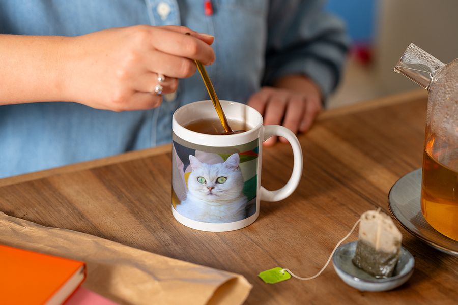 Alguien remueve con una cuchara una taza personalizada con la foto de un gato blanco mientras prepara una taza de té, ilustrando el consejo de Hofmann de completar un rincón de lectura hygge con una taza personalizada.
