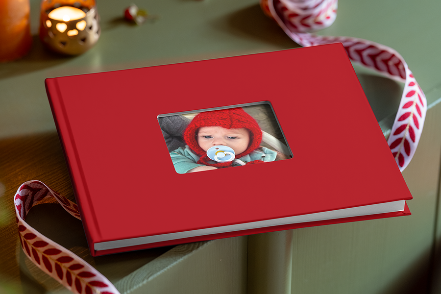 Álbum de fotos rojo con una ventana recortada en la portada, a través de la cual se ve un recién nacido con un gorro de punto rojo y un chupete, para ilustrar los consejos de Hofmann para crear un álbum de fotos con los primeros recuerdos del bebé.