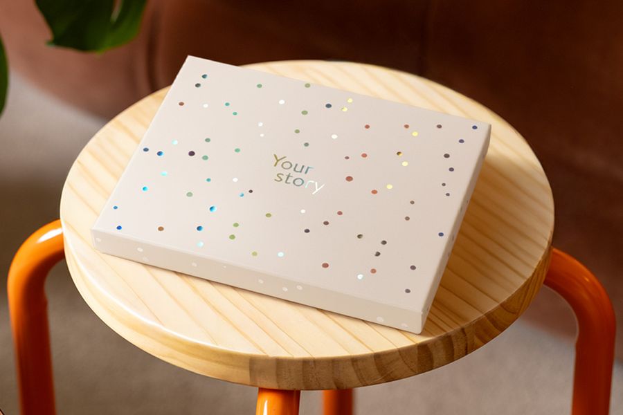 Una caja de presentación de Hofmann descansa sobre un taburete de madera, decorada con círculos holográficos plateados y el texto 'Your Story', ilustrando el diseño mate marfil con un toque de brillo holográfico.